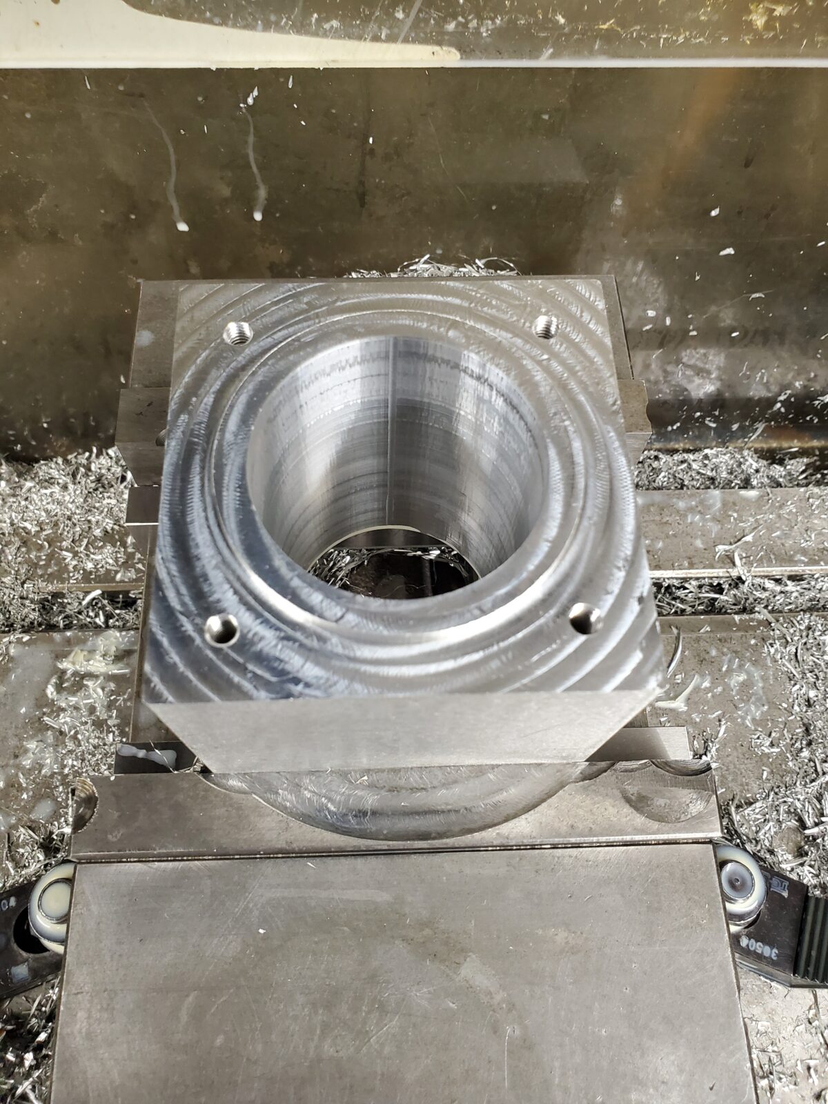 Aluminum block in a CNC mill vise with a deep milled bore and drilled and tapped holes, all machined in one setup