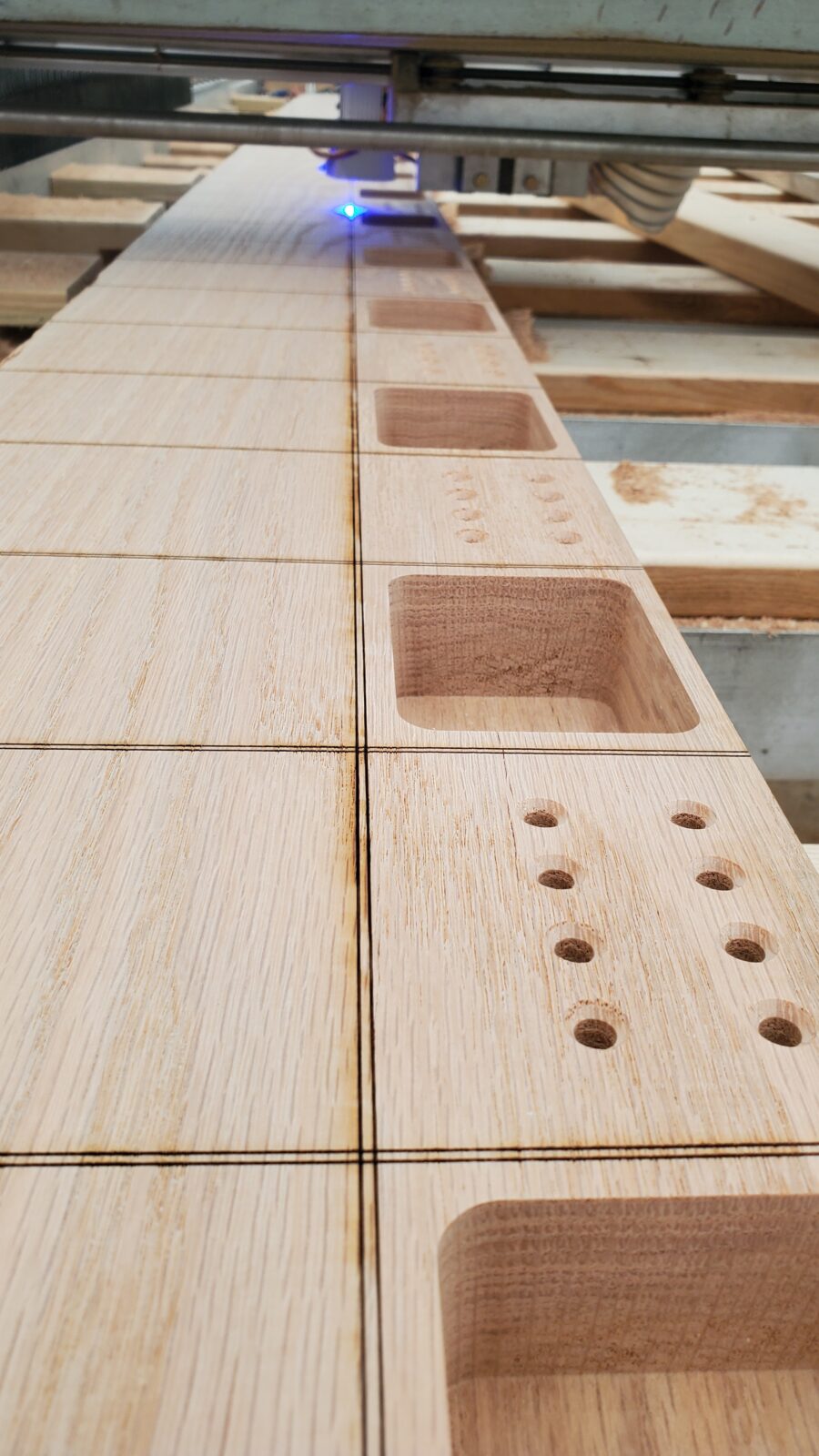 Red oak board on the CNC router with laser-engraved saw cut lines between machined box pockets and drilled holes, laser head glowing blue in the background