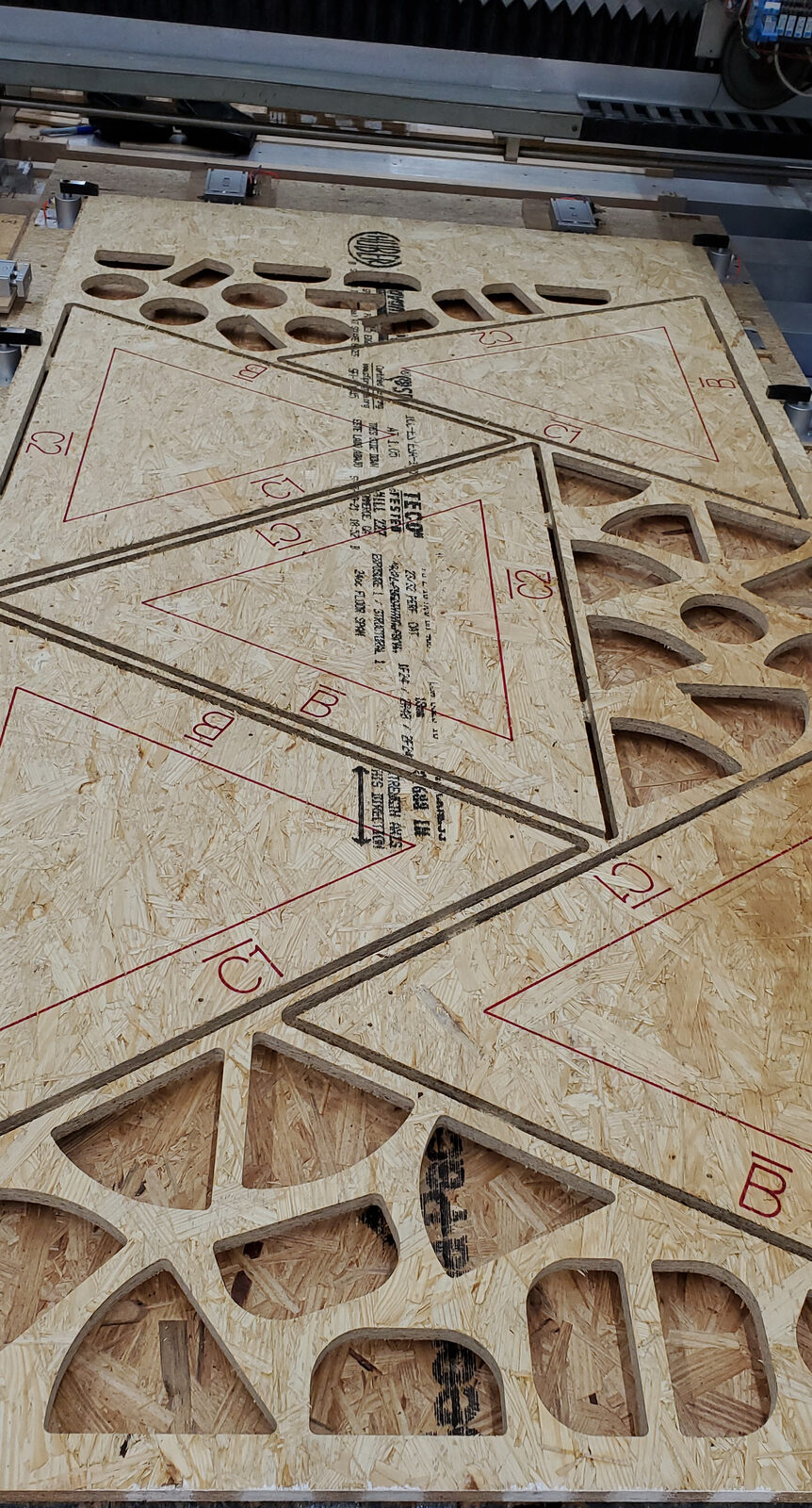 Engineered OSB subfloor sheet on a CNC router, marked with red part labels and outlines and nest-cut into triangles, with cutouts in the offcut